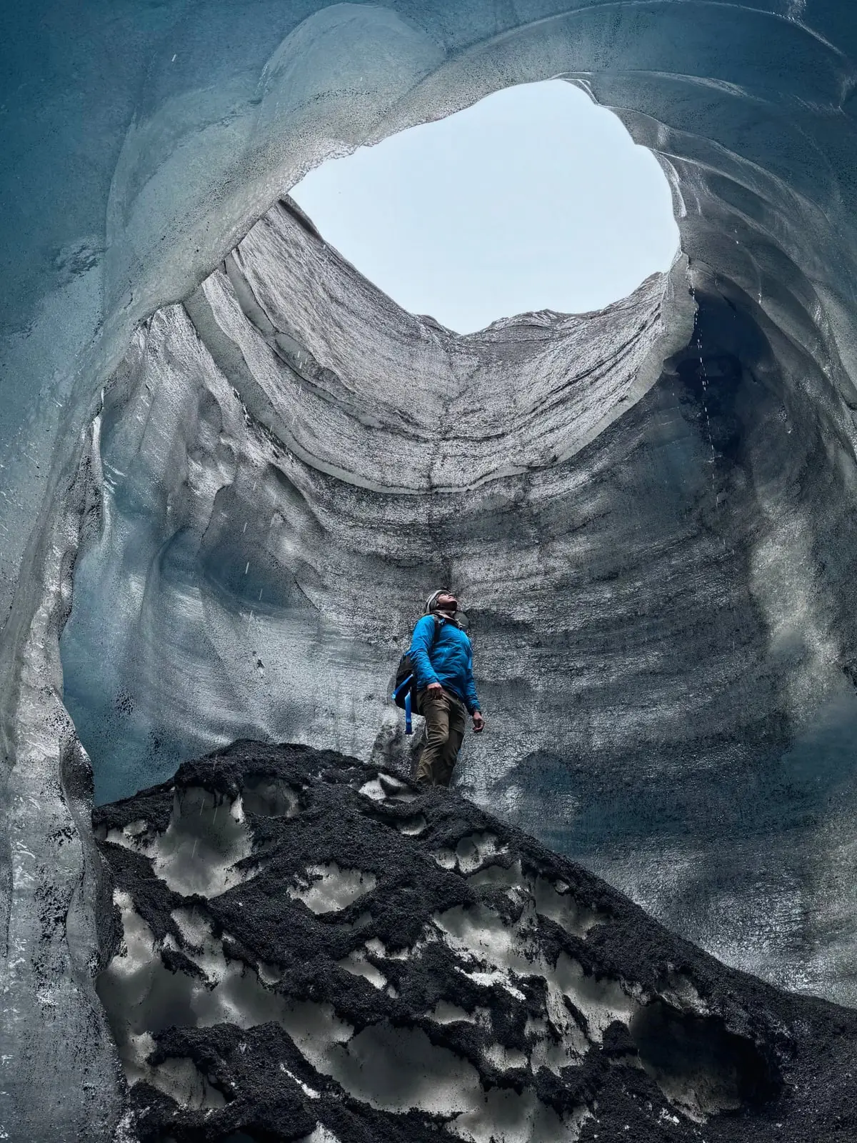 Alex N. Jose in a blue jacket, standing on dark rock inside an ice cave, looking up at an opening to the sky